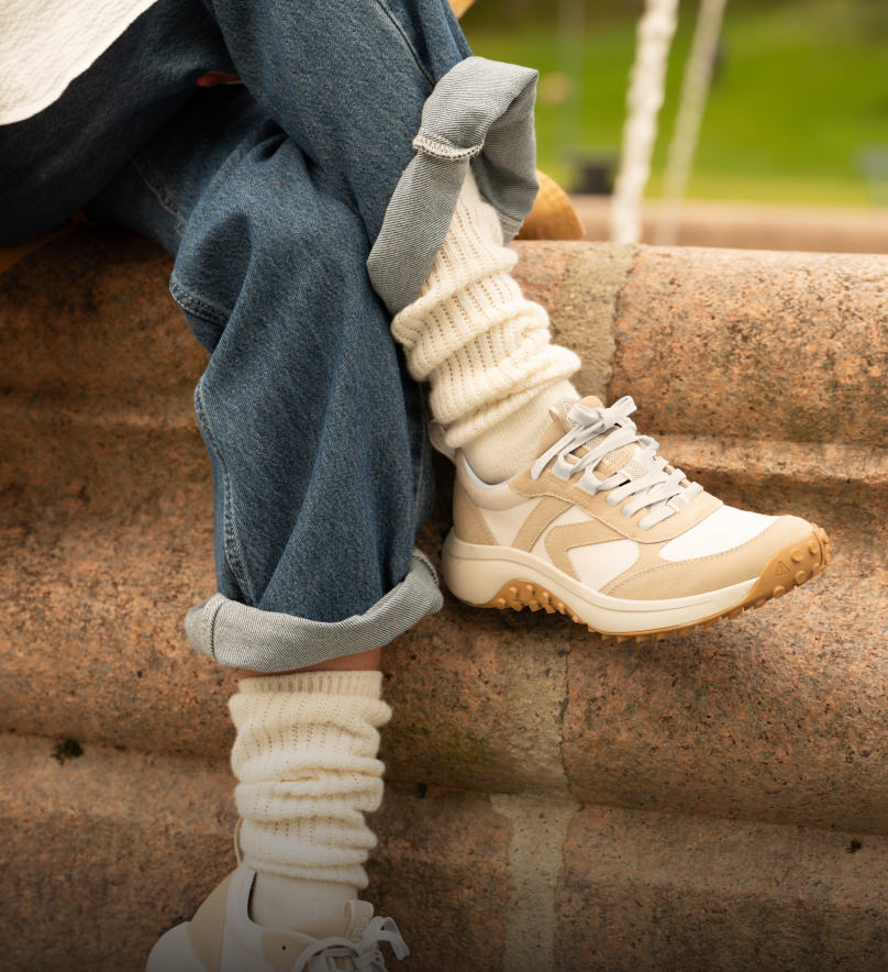 Waist down shot of woman in jeans and white socks  sitting on brick wall while crossing her legs and wearing tan and white KS86 sneakers