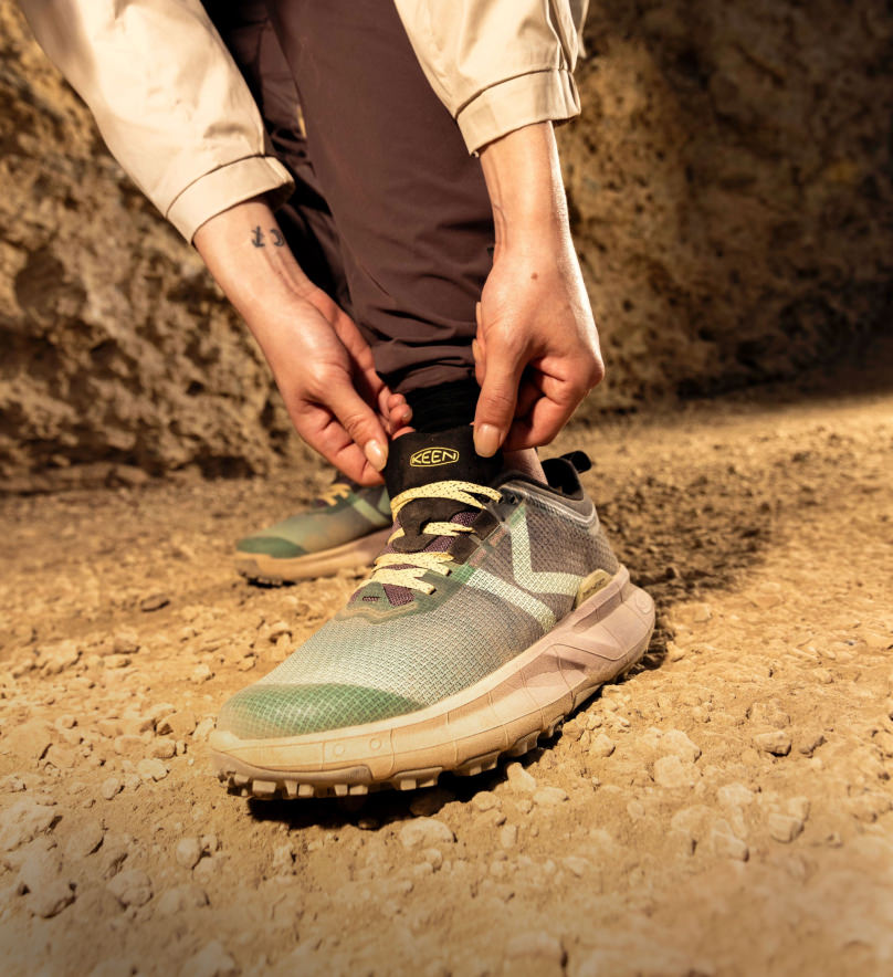 Knee down shot of man bending over to tie his blue 450 dirt sneakers on a trail. 